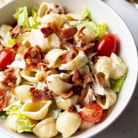 A close-up of a hearty BLT Chicken Pasta Salad, highlighting the tender pasta shells, golden grilled chicken pieces, bright green lettuce, and ripe cherry tomatoes, all coated in a smooth, light mayo dressing.