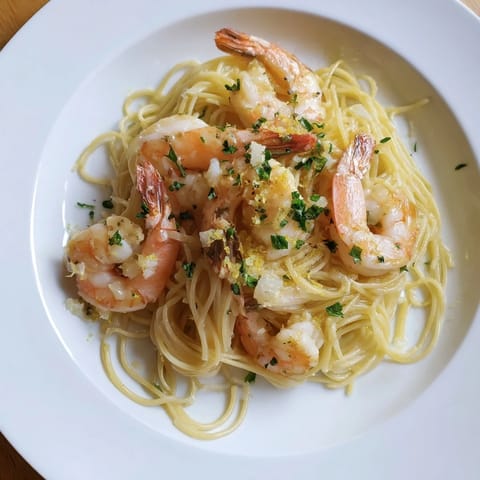 A close-up of succulent sautéed shrimp nestled in delicate angel hair pasta, bathed in a savory lemon butter sauce with flecks of garlic and red pepper flakes.